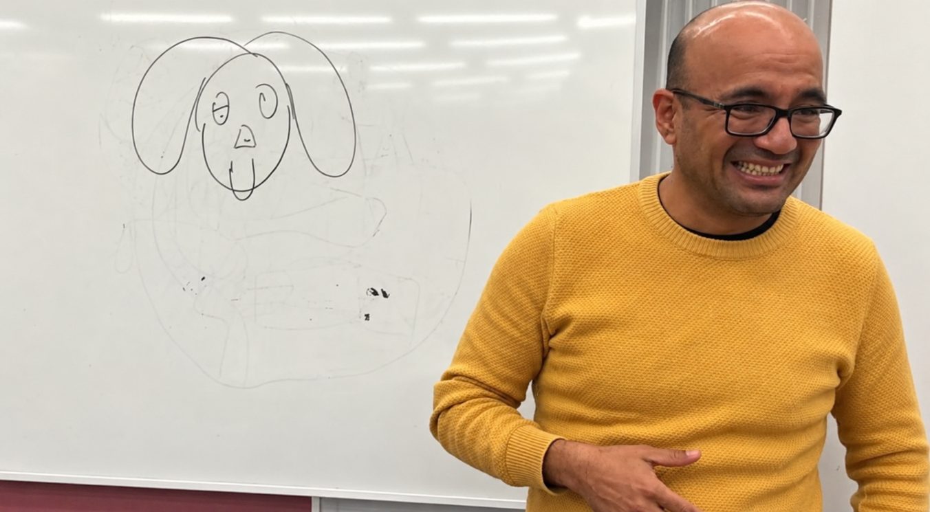 Omar Manky, riendo, junto a una pizarra con un perro dibujado. Nagoya, 2025.
